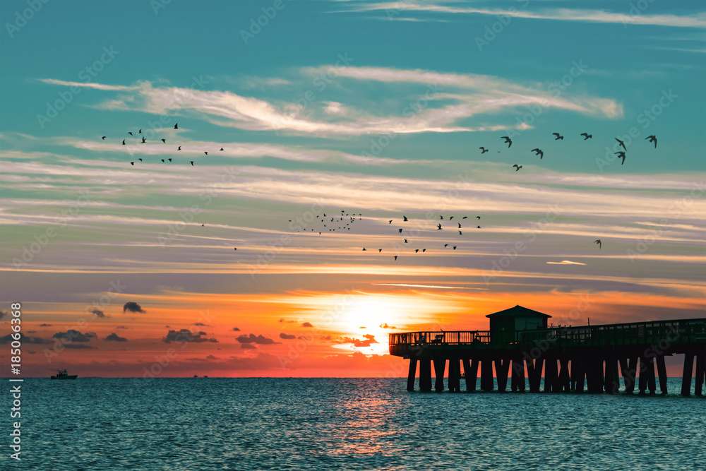 Fototapeta premium boats and birds passing the pier as the sun rises over the ocean