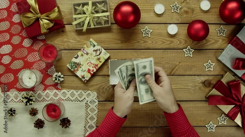 Man puts cash money into Xmas present box on wooden decorated table, top down shot