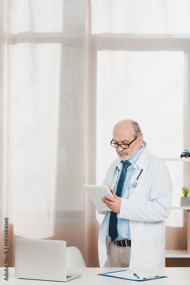 portrait of senior doctor in white coat with tablet in hands at workplace in clinic