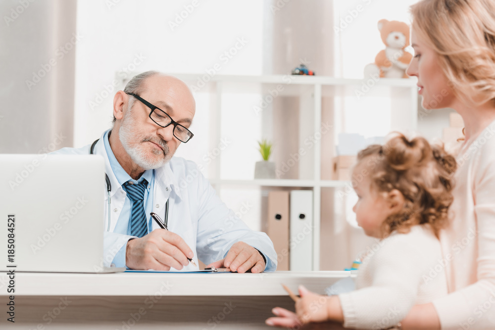 Fototapeta premium senior doctor making notes while mother and little daughter sitting at table near by in clinic