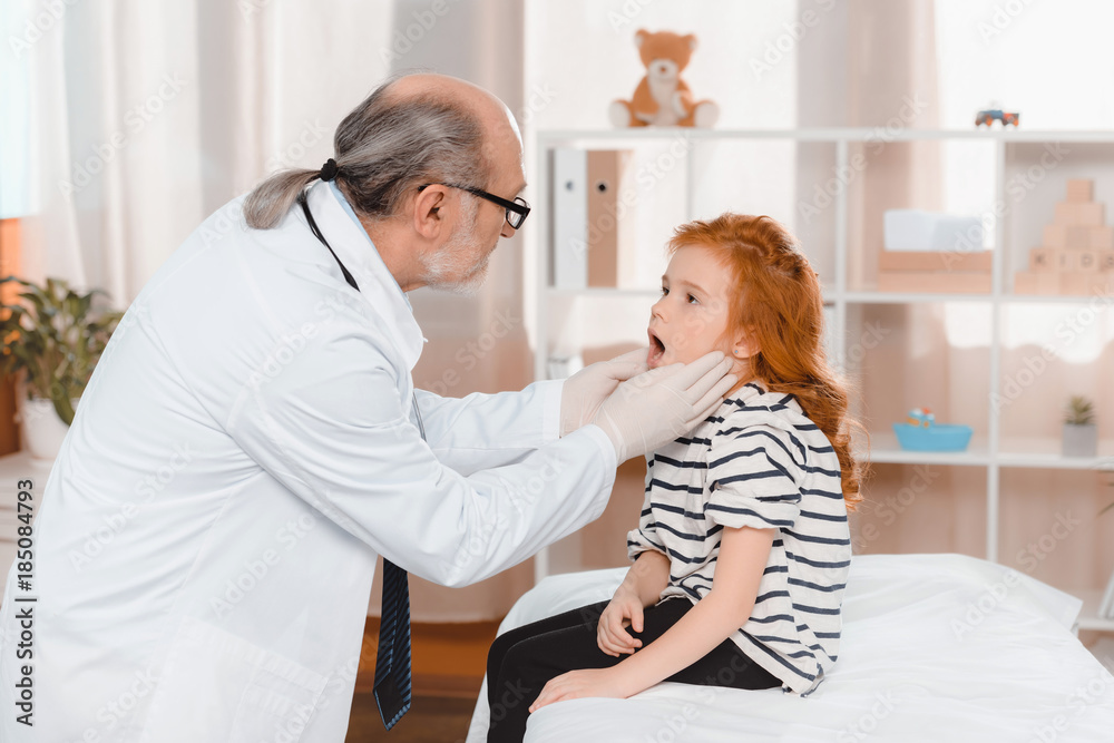 senior doctor in medical gloves examining little patients throat in clinic