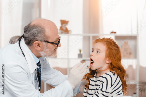 senior doctor in medical gloves examining little patients throat in clinic