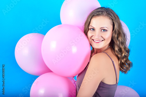 Happy smiling young woman having fun with pink helium air balloons over blue background. Girl with curly hair holding pink balloons. Beauty, holidays, birthday, valentine, fashion concept