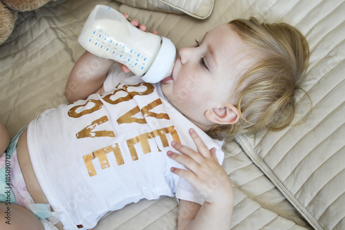 Маленький белый ребенок пьет молоко из бутылки лёжа на кравати. 