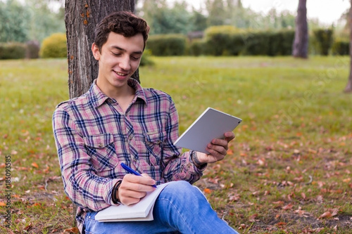 young men student sitting in the park next to the tree with the tablet in his hands .