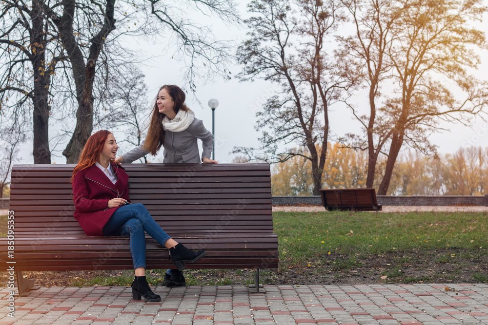 Fototapeta premium Female teenagers listening to music on smartphone at the bench in an autumn city park