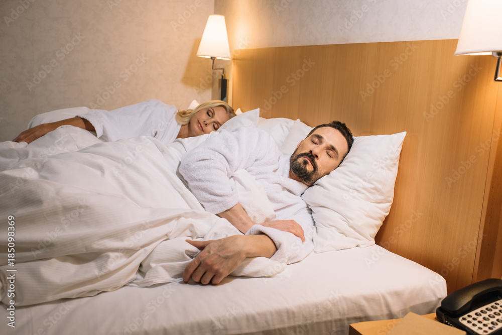 beautiful middle aged couple in bathrobes sleeping together in bed in hotel room beautiful middle aged couple in bathrobes sleeping together in bed in hotel room
