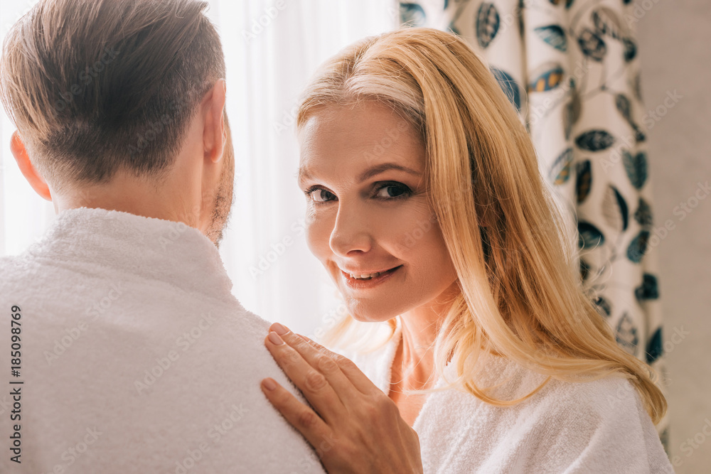 beautiful mature woman smiling at camera while embracing husband in bathrobe in hotel room beautiful mature woman smiling at camera while embracing husband in bathrobe in hotel room