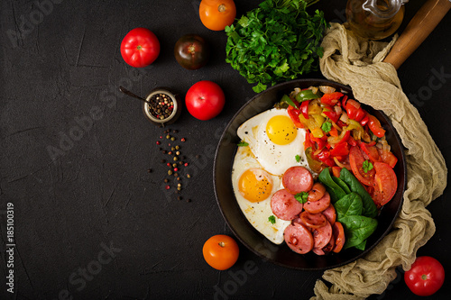 Breakfast. Fried eggs with sausage and vegetables in a frying pan on a black background in rustic style. Top view