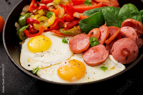 Breakfast. Fried eggs with sausage and vegetables in a frying pan on a black background in rustic style.