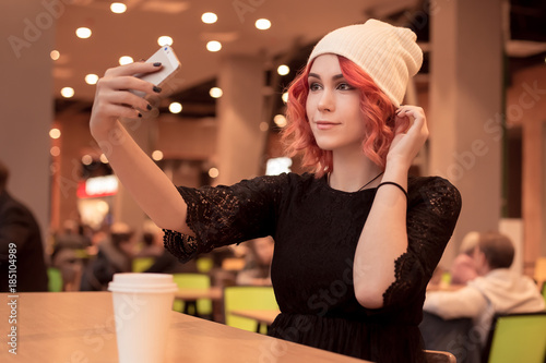 Beautiful girl with orange hair, in a cap photographes herself on a smartphone. Selfie on the phone. Sitting to wait friends alone at a food court in a cafe. Fashionable image. People Z. photo