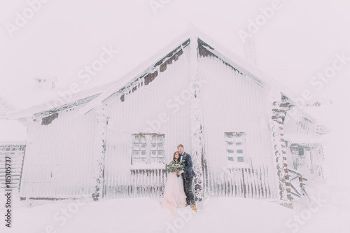 Young pretty pair of lovers. Winter. Date. Man hugging woman near the wooden house in mountain