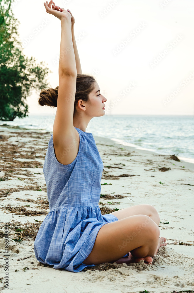 Girl is engaged in yoga on the beach. Young woman in a blue dress doing  stretching exercises outdoors Stock Photo