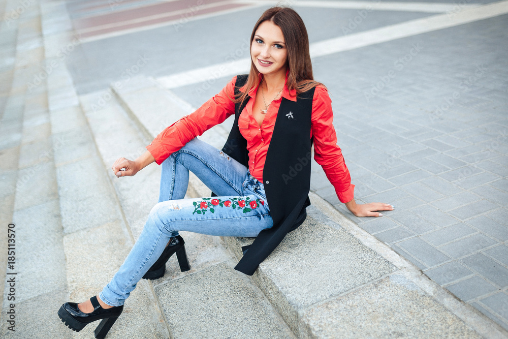 Young stylish woman in jeans and red shirt and a long sleeveless jacket  resting on stairs. Attractive girl with long hair sitting on steps. Stock  Photo