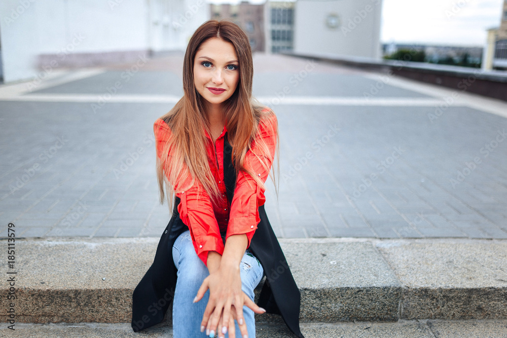 Young stylish woman in jeans and red shirt and a long sleeveless jacket resting on stairs. Attractive girl with long hair sitting on steps. Young stylish woman in jeans and red shirt and a long sleeveless jacket resting on stairs. Attractive girl with long hair sitting on steps.