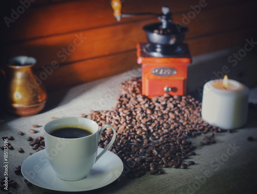 coffee beans on a wooden table, next to a cup of hot coffee, a hand grinder and a candle
