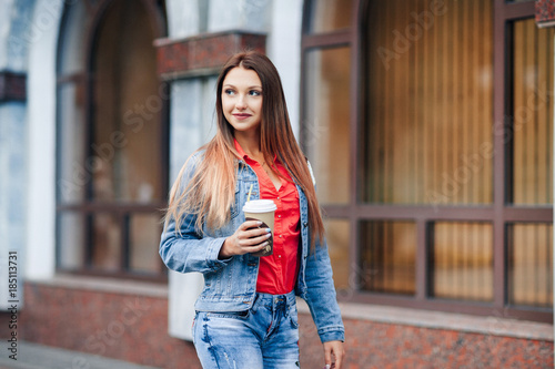 Portrait of young happy stylish woman in blue jeans and red shirt walking in the city centre and drinking coffee.