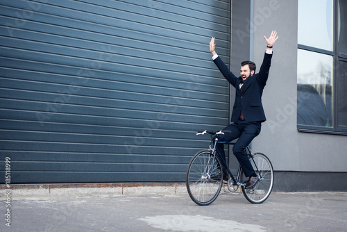 excited young businessman with outstretched arms riding bicycle on street