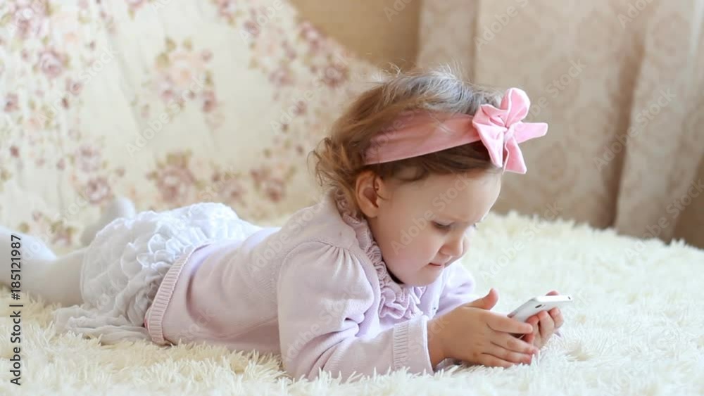 Child Looks cartoons and plays downloaded application on a smart phone close-up. A little cute funny girl lies in bed under the blanket and looks at the white phone screen. Child Looks cartoons and plays downloaded application on a smart phone close-up. A little cute funny girl lies in bed under the blanket and looks at the white phone screen.
