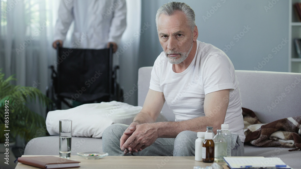 Sad man sitting on couch and looking into camera, male nurse bringing wheelchair Sad man sitting on couch and looking into camera, male nurse bringing wheelchair
