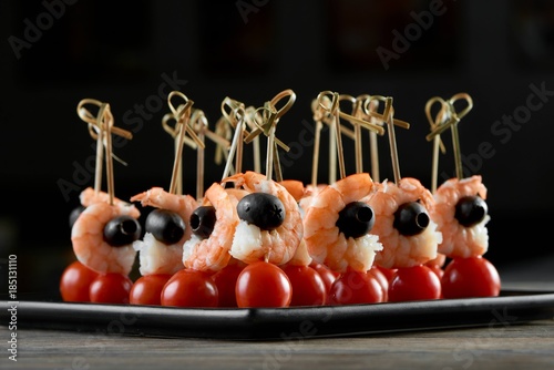 Close-up of tasty restaurant appetizer with shrimps,black olives and fresh red tomatoes. Delicious dish for light alcohol buffet or shampagne catering. Photo was taken on the black background