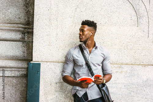 Modern College Student in New York. Wearing gray shirt, wristwatch, carrying shoulder leather bag, an African American guy standing by wall on street, listening music with earphone, reading red book..