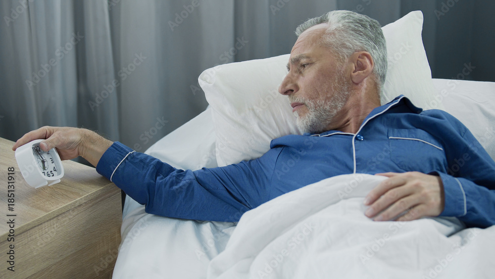 Obraz premium Elderly man hearing alarm clock, reluctant to wake up, lack of sleep and energy