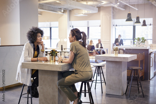 Businesswomen talking while sitting in cafeteria at creative office with colleagues in background photo