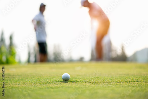Golf ball approach to the hold on the green. Couple golf player putting golf ball in the background. Lifestyle Concept.