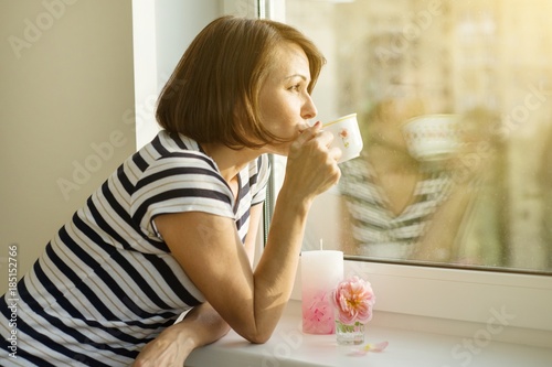 Adult woman drinks coffee, looks out the window at home.