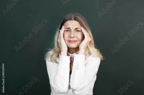 Picture of attractive blonde elderly woman suffering from migraine, being nervous about small things, having mournful look, squeezing her temples. People, age, stress, problems and sickness concept