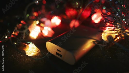 Silver powerbank external battery for fast charging smartphone or tablet pc lying on the table in the dark surrounded by flashing garland lights. photo