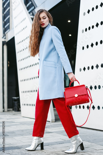 Outdoor full body portrait of young beautiful fashionable woman walking in street. Model wearing light blue coat, red pants, silvery ankle boots, holding trapeze handbag. Female fashion concept