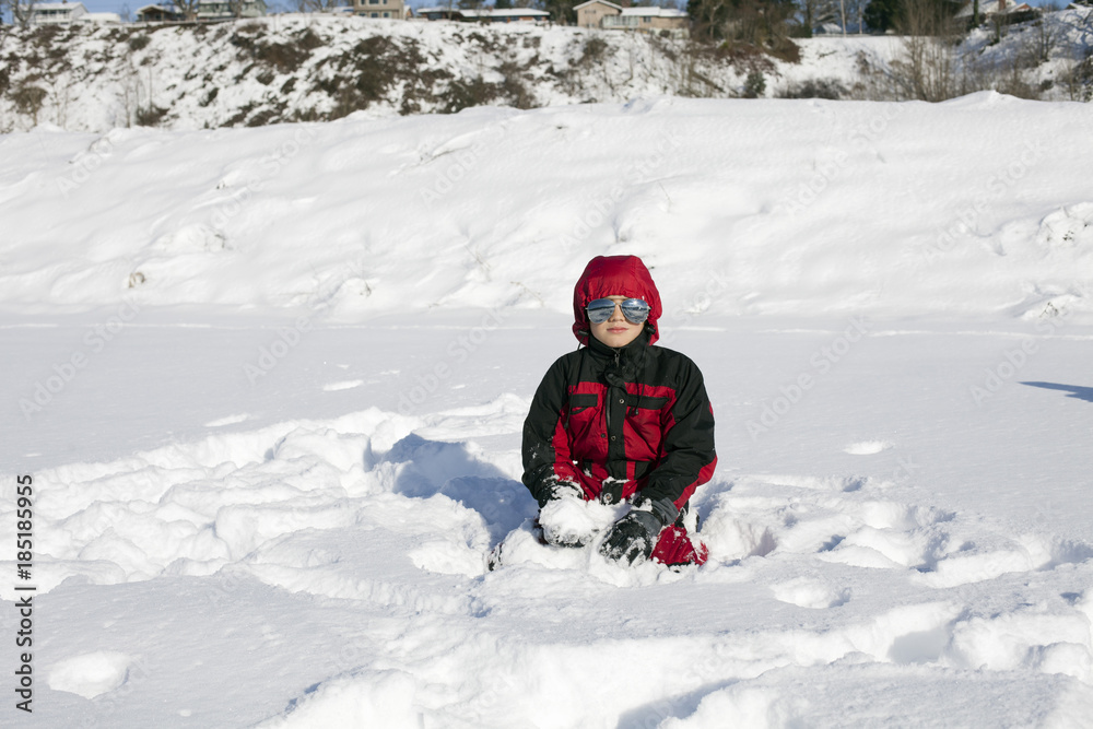 Boy in red snowsuit and reflective sunglasses sits in the snow Boy in red snowsuit and reflective sunglasses sits in the snow