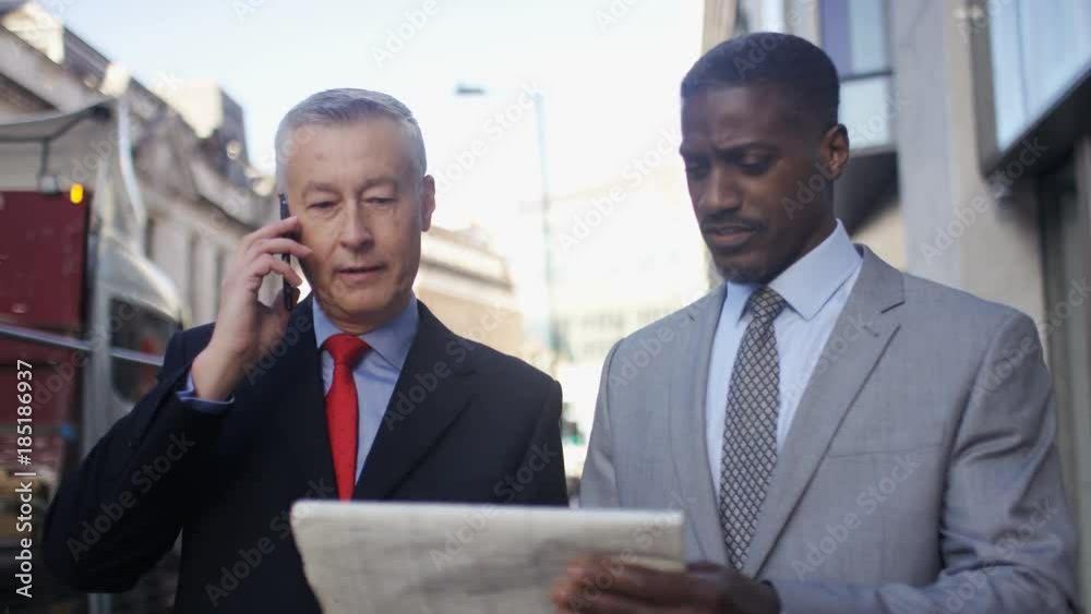 Two male investors talking on the phone as they walk and check share prices Two male investors talking on the phone as they walk and check share prices