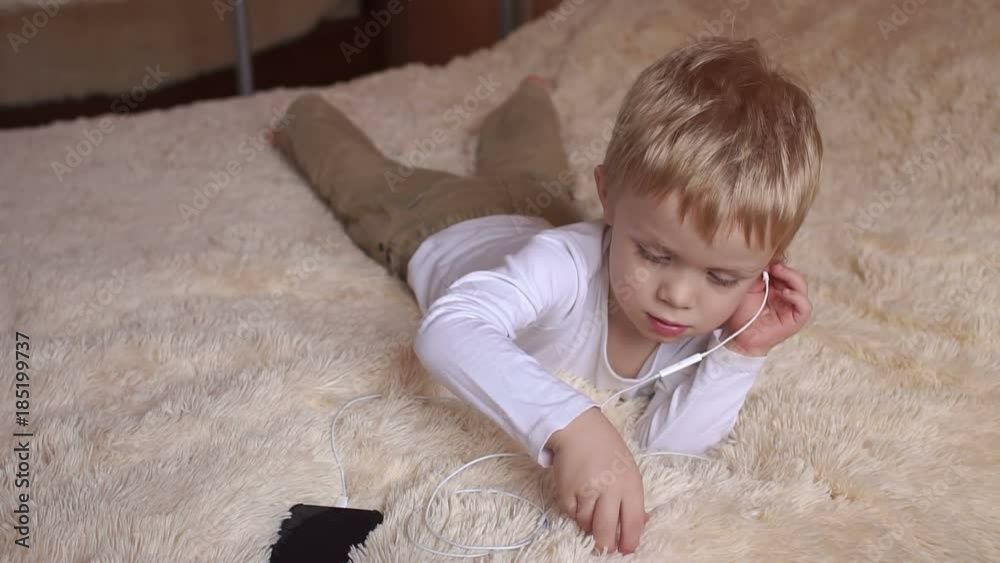 A little boy with blond hair laying on the bed on a fluffy blanket with headphones and listens to music. A little boy with blond hair laying on the bed on a fluffy blanket with headphones and listens to music.