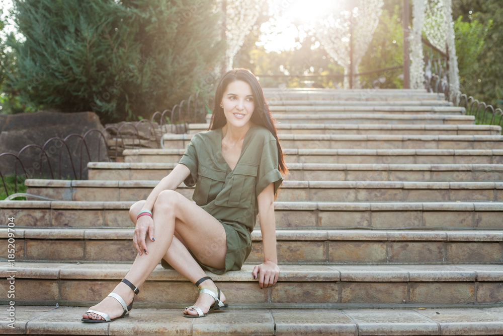Young attractive woman in green dress sitting on stone stairs in nature and smiling in sunlight. Concept of modern healthy woman Young attractive woman in green dress sitting on stone stairs in nature and smiling in sunlight. Concept of modern healthy woman