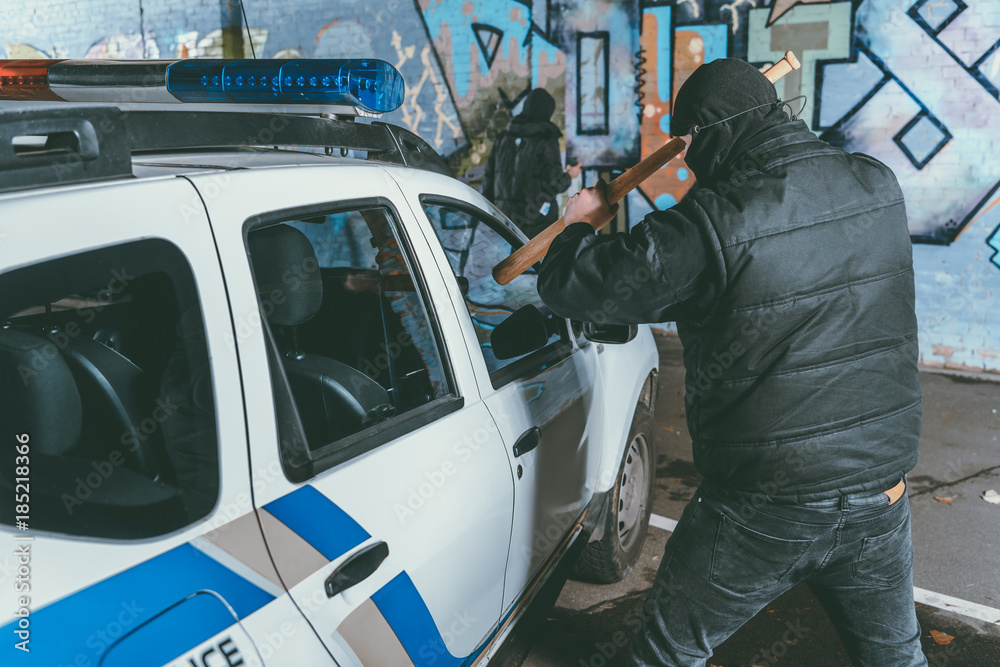 vandal crashing police car with baseball bat while another man painting graffiti on wall