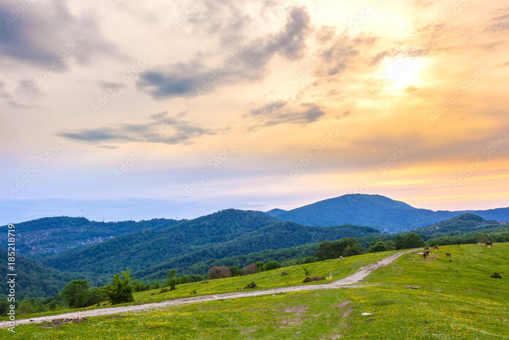 Sunset behind the clouds over the mountainous countryside, the road is in the green pastures. Rural evening spring landscape in Sochi, Russia.