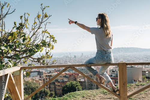 Rear view of young woman wearing in stripped t-shirt sitting on high point and looking at cityscape, pointing at something. Summer sunny day, rear view, bird's eye view of city, cityscape, horizon.