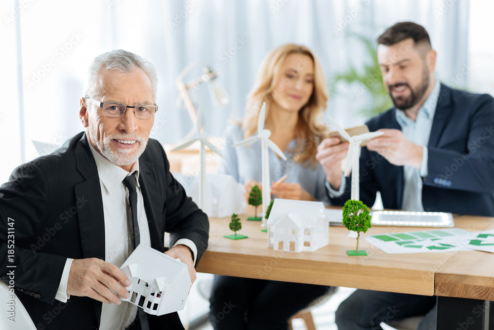 Successful project. Pleasant cheerful aged man sitting at the table with a lovely little house in his hands while his friendly colleagues sitting not far from him Successful project. Pleasant cheerful aged man sitting at the table with a lovely little house in his hands while his friendly colleagues sitting not far from him
