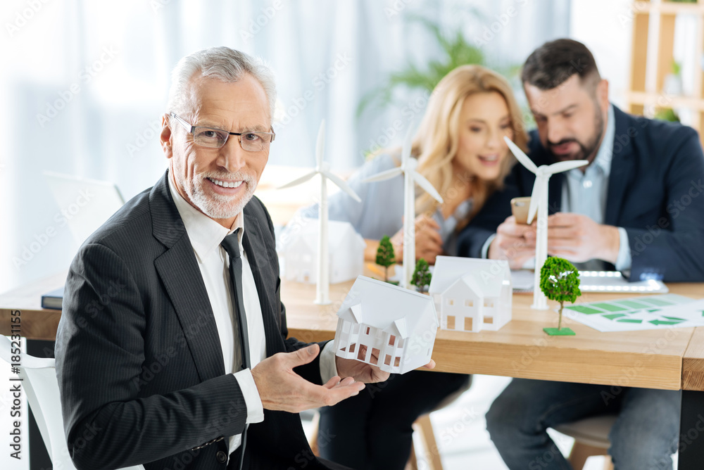 Important building. Smart excited aged man showing a lovely little while house while his skilled creative colleagues sitting at the table and discussing the construction of windmill turbines Important building. Smart excited aged man showing a lovely little while house while his skilled creative colleagues sitting at the table and discussing the construction of windmill turbines