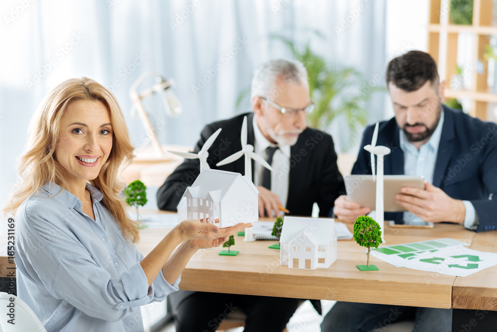 Cheerful woman. Cute emotional friendly woman looking glad and showing a nice model of a house while her enthusiastic colleagues sitting by her side and working at the progressive windmill turbines Cheerful woman. Cute emotional friendly woman looking glad and showing a nice model of a house while her enthusiastic colleagues sitting by her side and working at the progressive windmill turbines