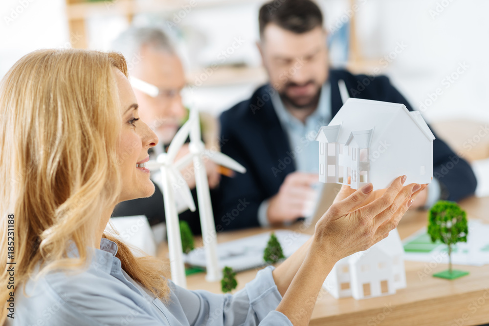 Lovely idea. Happy experienced clever engineer sitting next to her colleagues and admiring a lovely little model of an eco friendly house while holding it in her hands Lovely idea. Happy experienced clever engineer sitting next to her colleagues and admiring a lovely little model of an eco friendly house while holding it in her hands