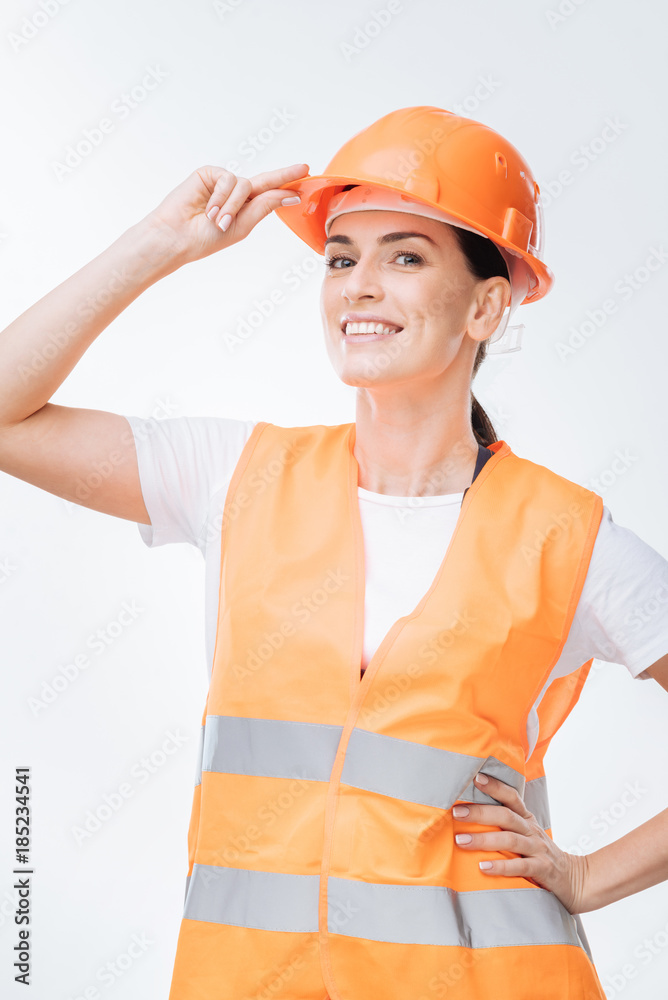 Future perspective. Merry glad female worker standing on the isolated background while fixing hard hat and laughing 