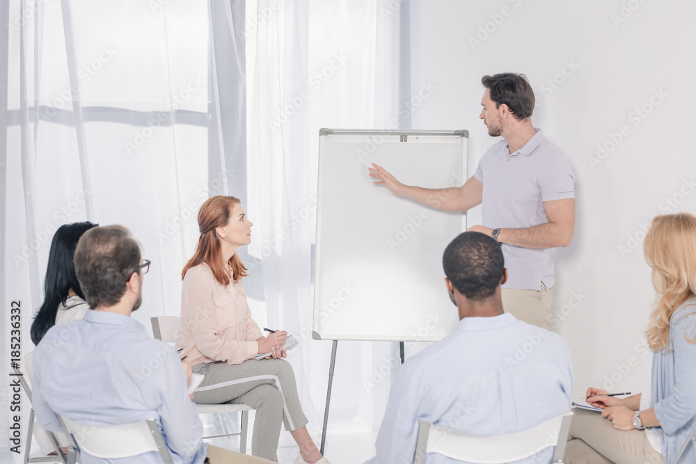 Obraz premium psychotherapist pointing at blank whiteboard and multiethnic group sitting on chairs during therapy
