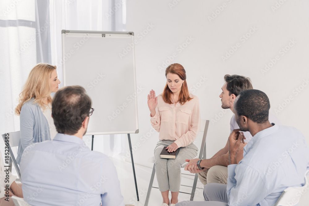 woman with closed eyes holding hand on Holy Bible while sitting with multiethnic people during group therapy woman with closed eyes holding hand on Holy Bible while sitting with multiethnic people during group therapy