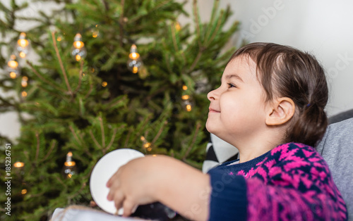 Happy holidays and Merry Christmas! Side view of cute little toddler girl with christmas present. Kid enjoy the holiday and open the gift sitting on grey sofa in the room. Copyspace