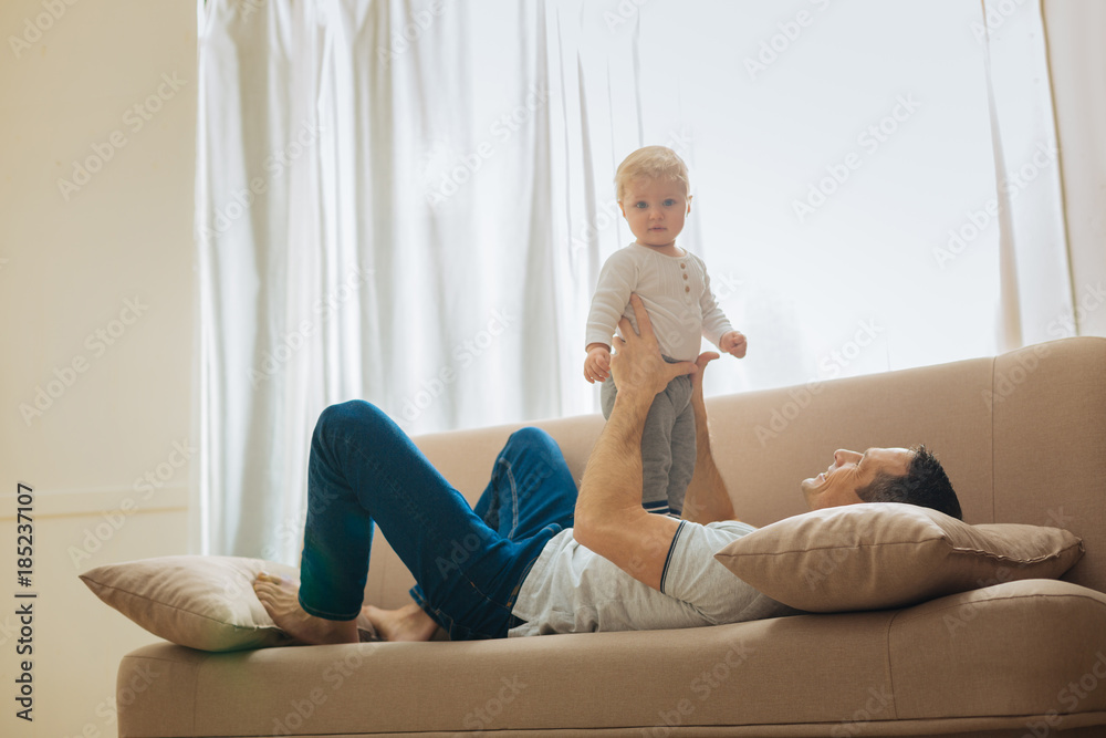 Relax. Kind loving father relaxing on a soft big sofa and looking at his adorable little child standing on his tummy and feeling curious Relax. Kind loving father relaxing on a soft big sofa and looking at his adorable little child standing on his tummy and feeling curious
