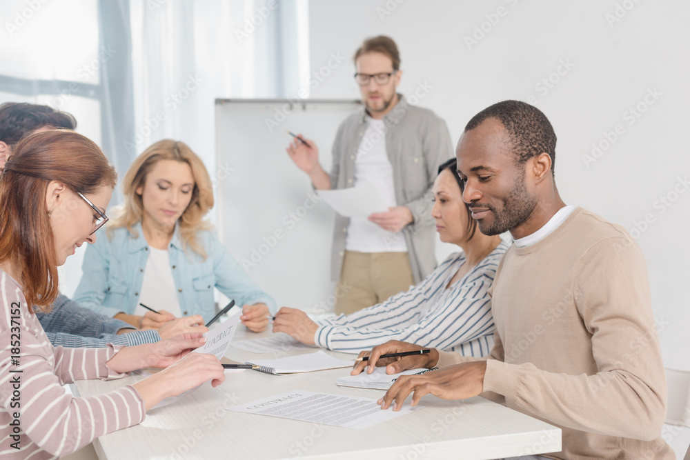 multiethnic team working with papers together while man standing near whiteboard behind multiethnic team working with papers together while man standing near whiteboard behind